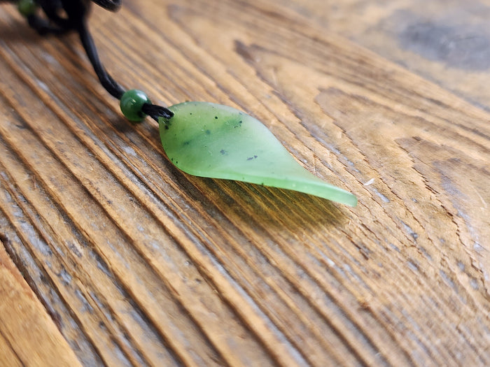 Single pale green jade leaf pendant on a black cord with a green bead on a wooden surface