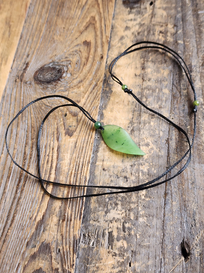 Jade leaf pendant on a black cord with a green bead resting on a rustic wooden surface