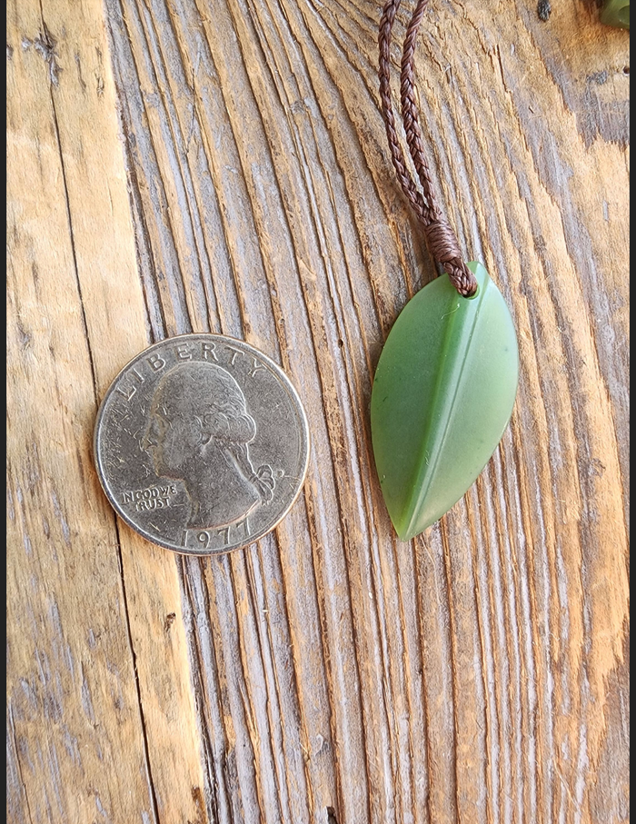 Green jade leaf pendant on brown braided cord beside a 1971 quarter for scale