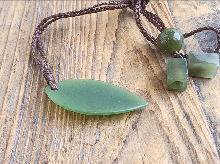Jade leaf pendant with a round bead and two square beads on a brown braided cord against a wooden surface