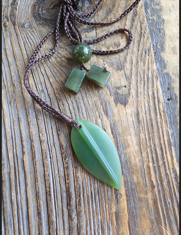 Green jade leaf pendant on a brown braided cord accompanied by a round bead and two cube beads on a rustic wood background