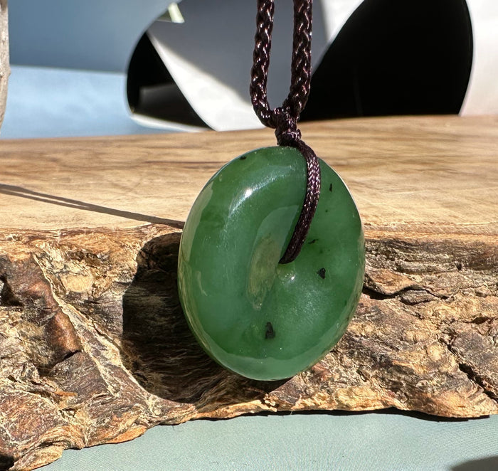 Green jade donut pendant on a brown braided cord resting on a rustic log.