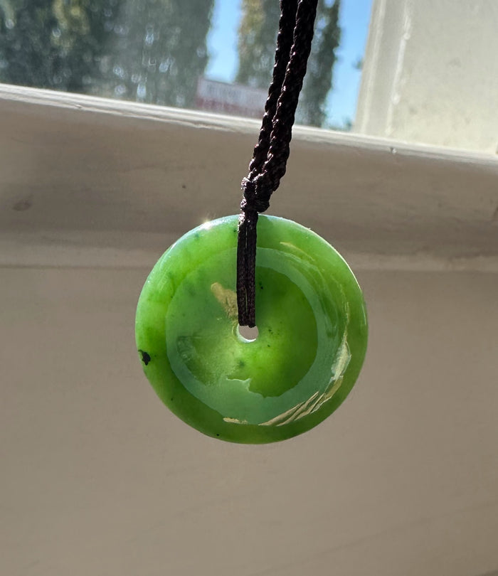 Green jade donut pendant on a dark brown braided cord hanging by a sunlit window.
