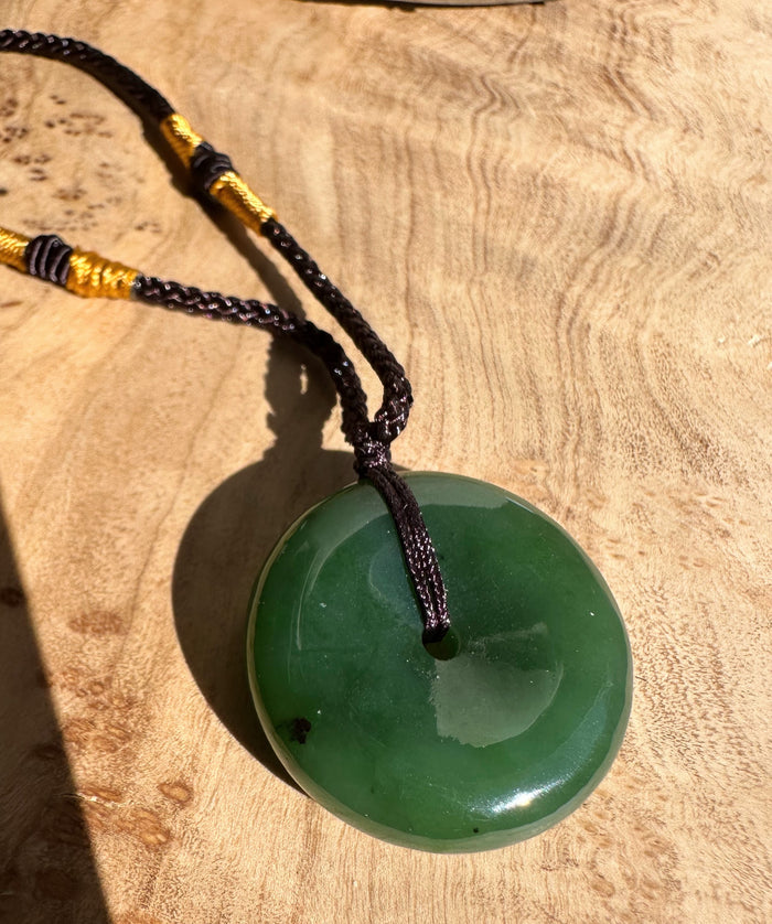 Green jade donut pendant on a braided cord with gold wrap placed on a wooden surface.