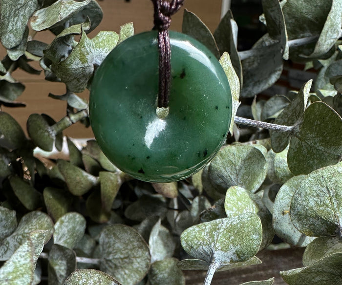 Green jade donut pendant threaded on a brown cord hanging among dusty green eucalyptus leaves