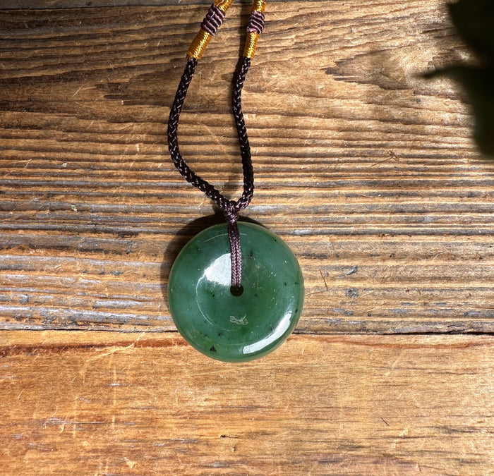 Green jade donut pendant threaded on a double brown braided cord with yellow wrapped accents.