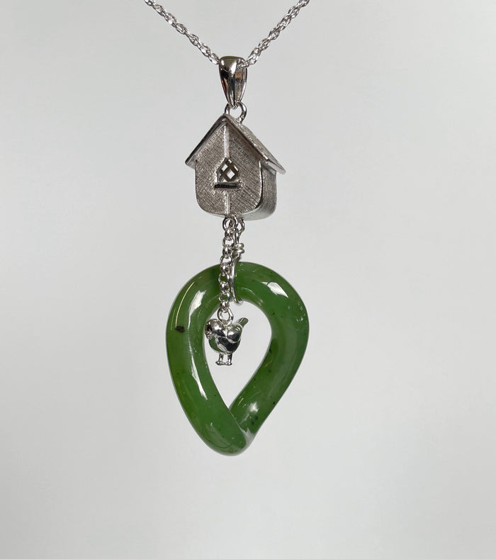 Silver birdhouse pendant with lattice window above a green jade loop pendant on a chain, with a tiny silver bird charm inside.