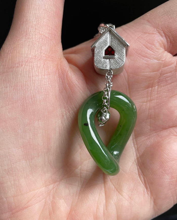 Jade green heart shaped loop pendant attached to a silver birdhouse charm on a chain held in a hand.