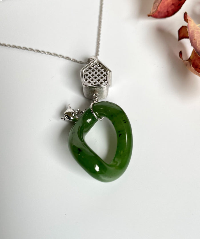 Green jade loop pendant attached to a silver birdhouse charm on a necklace.