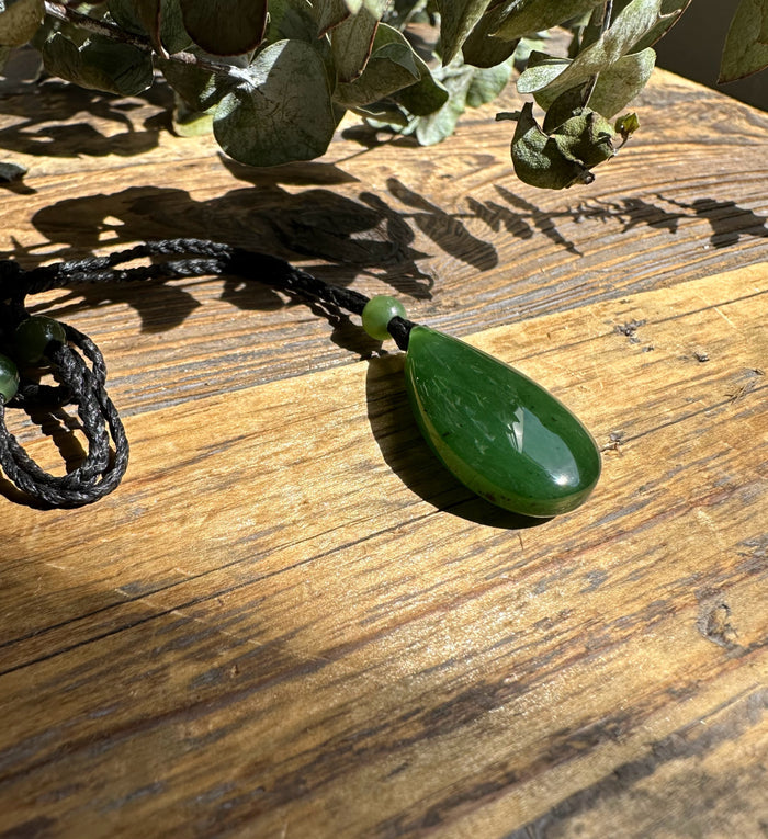 Green jade teardrop pendant on a black braided cord with a round bead above, displayed on a wooden surface