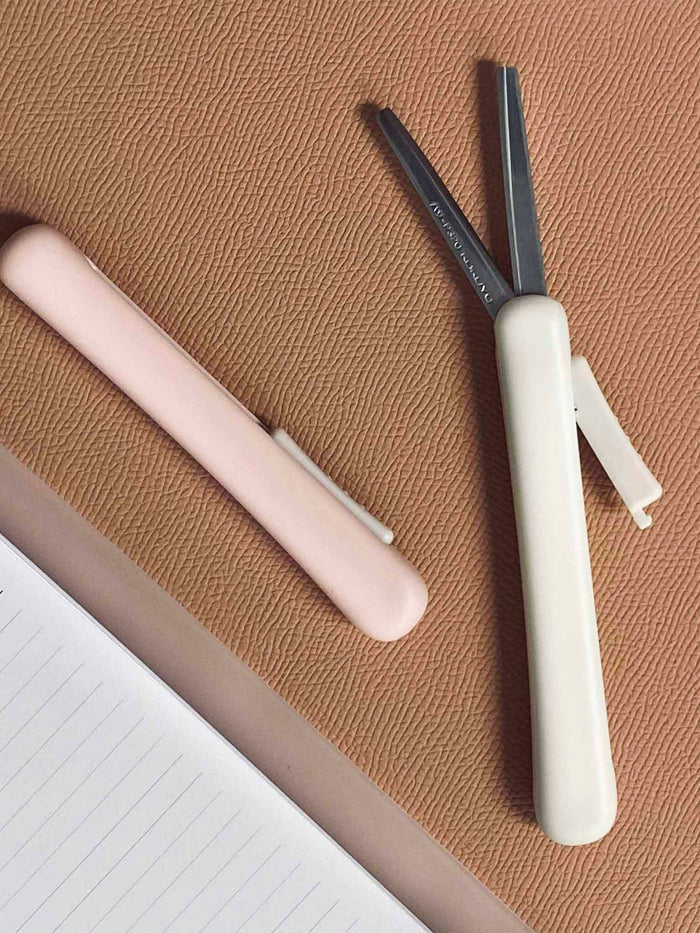 White and Peach scissors on a rust colored surface next to a planner. The white scissors are extended to show the blades.