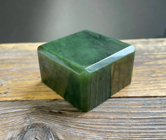 Polished deep green mineral specimen in a cuboid shape resting on a worn wooden surface.