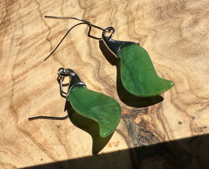 Green nephrite jade leaf earrings with silver caps and curved hooks resting on a wood surface.