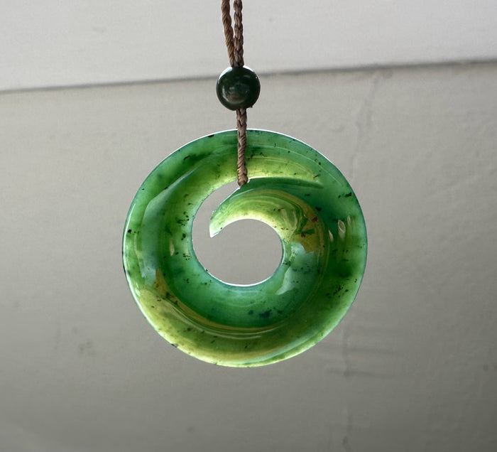 Circular green jade pendant with a large hole hanging from a brown braided cord with a small green bead above