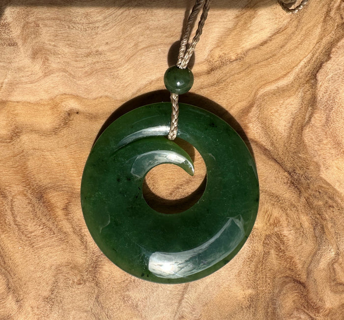 Green jade donut pendant with inner hole hanging from a braided cord against a wooden background.