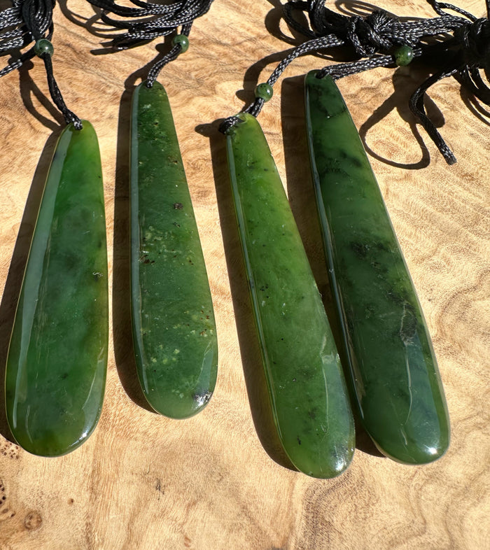 Four green jade drop pendants on black cords with small beads laid on a wooden surface