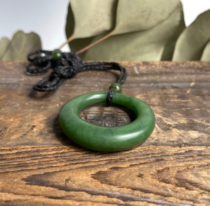 Round green jade donut pendant on a black braided cord against a rustic wooden surface.