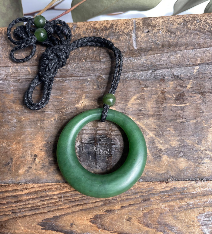 Close up of round green jade donut pendant on a black braided cord with green beads.