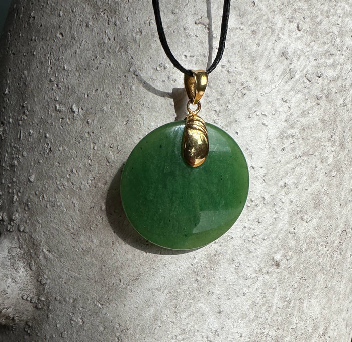 Round green jade pendant with gold bail on black cord against a concrete background