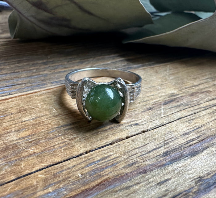 Silver ring on a wooden surface featuring a large green jade bead cradled by curved side settings.