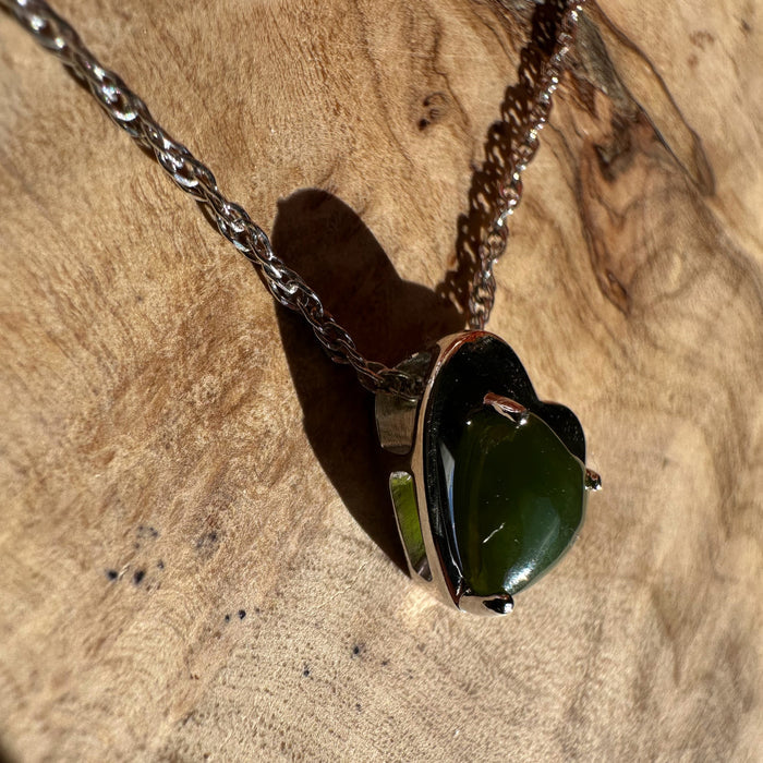 Silver heart pendant with a glossy green gemstone on a silver chain resting on a rocky surface