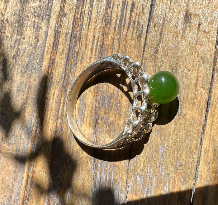 Silver ring with a round green jade cabochon and intricate openwork shoulders on a wooden surface.