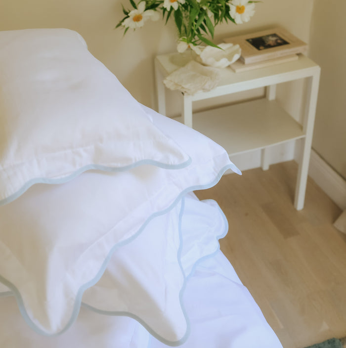 Stack of white pillows with blue trim on a bed with a nightstand in the background