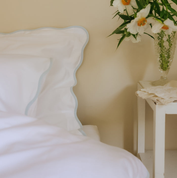 White pillow with blue trim on a bed with a vase of flowers on a nightstand in the background.